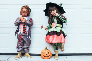Two girls dressed as witch and zombie eating candy celebrating Halloween at the garage door next to...