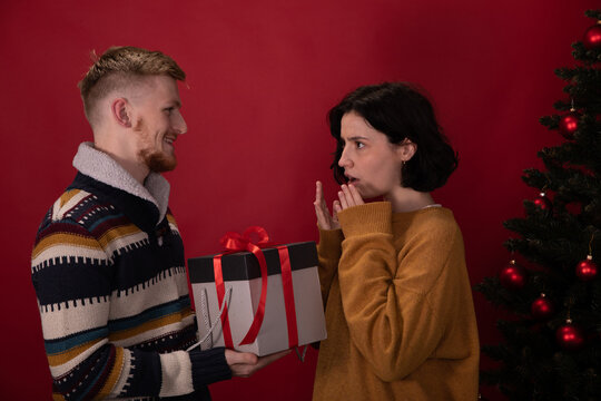 Young Couple Celebrating New Year At Home In Front Of Xmas Tree, Happy Man Presents Christmas Unpleasant Bad Gift Box To Upset Woman. Red Background, Winter Holidays, Unjustified Expectations Concept