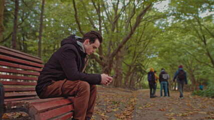 Fototapeta premium Young guy with a phone in his hands sitting on a bench in the park. In the background, a group of friends walking in the park. The concept of loneliness and dependence on the Internet and smartphones.