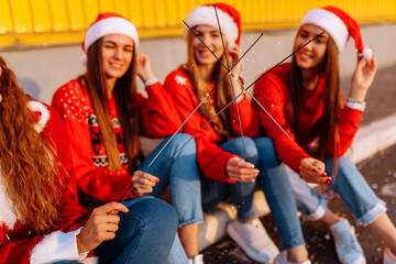 Group of happy young friends, girls in festive Santa Claus hats, holding sparklers, celebrating New Year, standing at the yellow wall outdoors