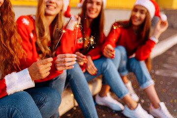 Company of cheerful young women celebrating the New Year in Santa Claus hats, with sparklers, outdoors