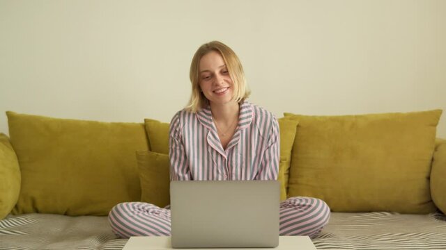 Girl in a pink striped pajama sitting on a yellow sofa in front of a laptop talking to friends over zoom or video call at home