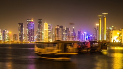 Beautifully illuminated high skyscrapers of Doha business city center at night and blurred ship (because of long exposition) sailing along the harbour. Qatar.