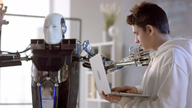 Medium closeup of humanoid android making moves programmed by high-school caucasian student typing on keyboard and smiling