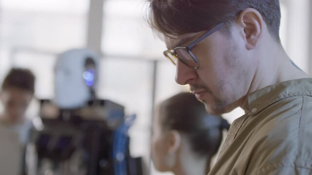 Medium closeup of male teacher typing programs for humanoid on laptop demonstrating its abilities to teenage mixed race students in bright classroom with panoramic windows