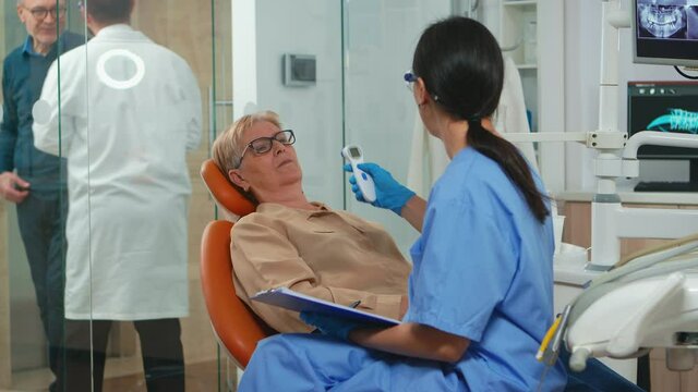 Nusre measuring old woman temperature before dental examination waiting for doctor. Dentist assistant interrogating senior woman and taking notes on clipboard sitting on stomatological chair