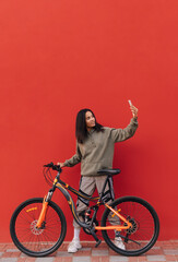 Positive woman sitting on a bicycle on a background of a red wall and taking a selfie on a smartphone. Isolated. Vertical