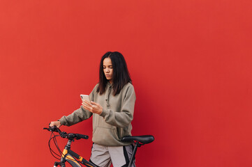 Pretty hispanic woman with bicycle in hand uses smartphone on red background. Beautiful woman with red bicycle isolated. Copy space