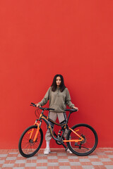 Fototapeta na wymiar Cheerful woman in sportswear stands with a bicycle on a background of a red wall and poses for the camera. Cycling down the street.
