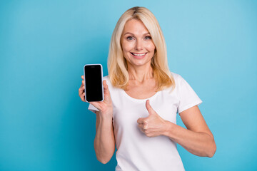 Positive old lady hold smartphone select thumb up sign wear white t-shirt isolated blue color...
