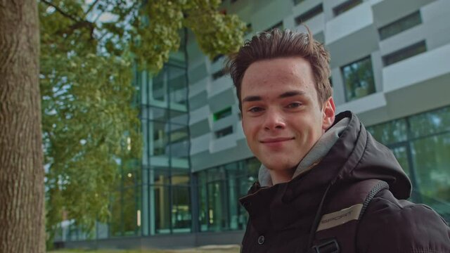 Handsome young man in modern warm clothes with a backpack on his shoulders looks back at the camera. Portrait of a successful and confident student. Teenager smiling at the camera.