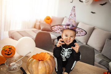 Halloween, holiday and childhood concept - Smiling little boy in party costume of skeleton with jack-o-lantern pumpkin sitting on table and having fun at home