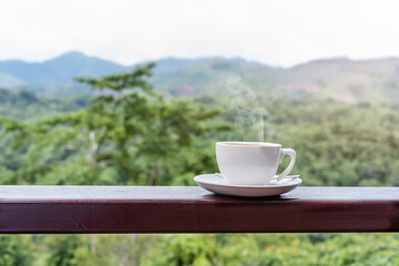 Close-up of a hot white coffee cup on wooden table with the scenic view of mountain in the morning sunlight and beautiful green nature background with copy space for text