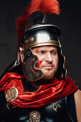 Headshot of bearded legionary he dressed in dark armour and helmet with red cape in dark background.