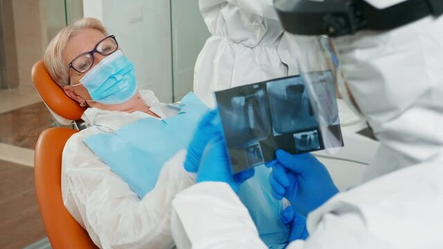 Dentist with face shield reviewing panoramic mouth x-ray image to patient during global pandemic. Assistant and doctor talking with senior woman wearing suit, coverall, protection suit, mask, gloves