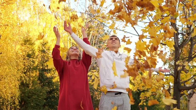 Positive young couple throw armfuls of dry bright yellow leaves and hugs in autumn park slow motion