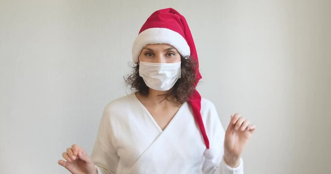 Young woman in medical mask and Santa Claus hat dancing and having fun. Close up of female in Christmas hat and protective mask.Concept of safe Christmas celebration during coronavirus pandemic