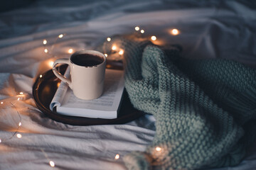 Cup of fresh hot coffee on open book staying on wooden tray with knitted textile in bed close up....