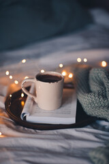 Cup of fresh coffee on open folded book staying on wooden tray in bed close up. Good morning. Winter holiday season. Cozy home atmosphere.