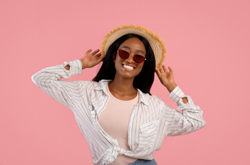 Portrait of gorgeous black woman in straw hat and sunglasses posing on pink studio background