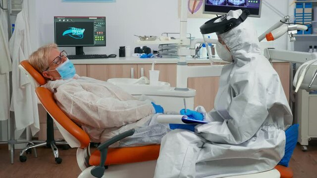 Nusre in protection suit measuring old woman temperature before dental examination during global pandemic. Assistant with face shield, mask, interrogating senior woman and taking notes on clipboard
