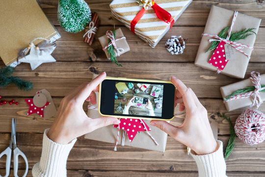 Flat lay of Woman s hands wrapping Christmas gift and taking pictures on the phone. Unprepared presents on wooden background with decor elements and items, top view. New year DIY packing Concept.