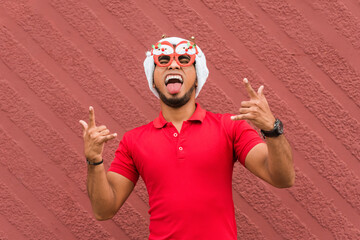 Young handsome  man with beard wearing casual shirt and glasses over red wall shouting with crazy expression doing rock symbol with hands up. Music star. Heavy music concept.