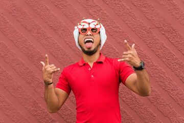 Young handsome man with beard wearing casual shirt and glasses over red wall shouting with crazy expression doing rock symbol with hands up. Music star. Heavy music concept.
