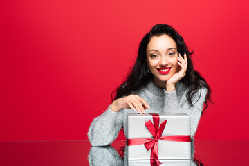 Fototapeta premium young happy woman in sweater looking at camera near wrapped present isolated on red
