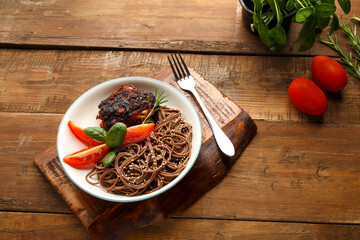 A plate of soba with chicken tomatoes and spinach on a wooden stand and a fork near the spinach and tomatoes on the table.