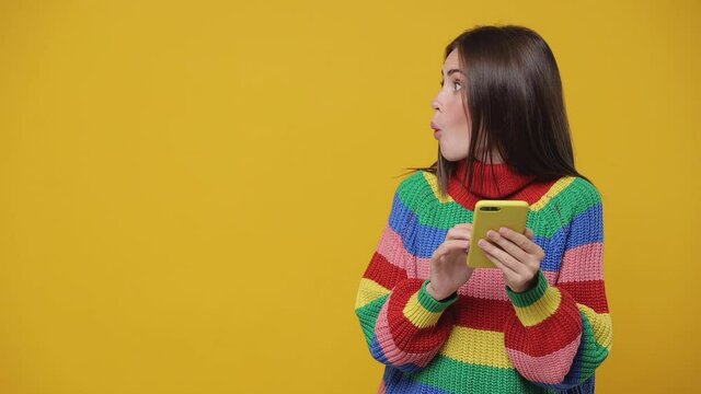Shocked young woman 20s years old in colorful sweater isolated on yellow background studio. People lifestyle concept. Using mobile phone typing sms mesage looking aside put hand on head chest say wow
