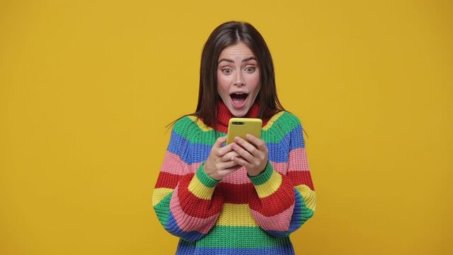 Surprised young woman 20s years old in colorful sweater isolated on yellow background studio. People lifestyle concept. Using point finger on mobile cell phone typing sms mesage say wow spreading hand