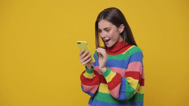 Shocked amazed young woman 20s years old in colorful sweater isolated on yellow background studio. People lifestyle concept. Using mobile cell phone typing sms mesage browsing say wow put hand on head