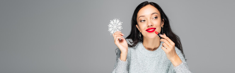 pleased woman with red lips holding decorative snowflake and applying lipstick isolated on grey, banner