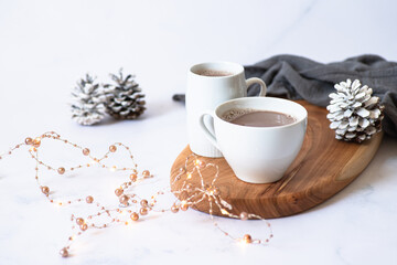 Obraz na płótnie Canvas Two white cups with hot chocolate or cocoa drink on a wooden board on a light marble background with festive winter decorations. Winter mood. Frontal view. Copy space. Selective focus