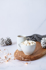 Obraz na płótnie Canvas Two white cups with hot chocolate or cocoa drink with marshmallows on a wooden board on a light marble background with festive winter decorations. Winter mood. Close up. Selective focus 
