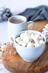 Two white cups with hot chocolate or cocoa drink with marshmallows on a wooden board on a light marble background with festive winter decorations. Winter mood. Close up. Selective focus 