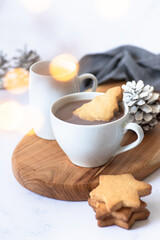 Two white cups with hot chocolate or cocoa drink with Christmas tree shape cookie on a wooden board and light marble background with festive winter decorations. Winter mood. Close up. Selective focus 