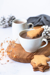 Two white cups with hot chocolate or cocoa drink with Christmas tree shape cookie on a wooden board and light marble background with festive winter decorations. Winter mood. Close up. Selective focus 