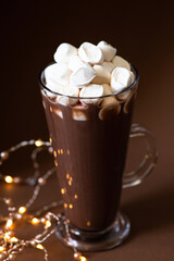 Cocoa drink with marshmallows in a glass on a brown background with festive winter decorations. Winter mood. Minimalistic monochrome composition. Selective focus. Frontal view. Vertical. Copy space