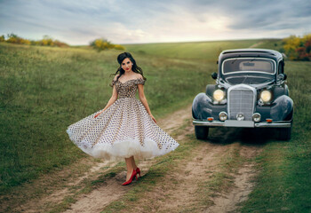 Young beautiful happy retro woman near the old car. Attractive elegant lady in vintage white polka black dot retro dress. Pin-up hairstyle. Cheerful girl standing on road. Autumn nature, yellow trees