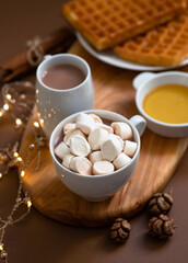 Winter sweet breakfast with cocoa drink with marshmallows, waffles, honey, served on a wooden board and a brown background with festive Christmas decorations. Winter mood.  Close-up. Selective focus 