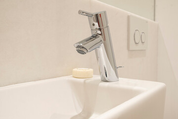 Close-up of a metal faucet with a white sink in a bathroom tiled with light tiles