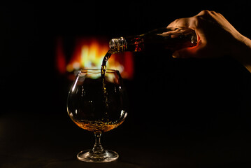 Woman pours cognac into a glass on the background of the fireplace.
