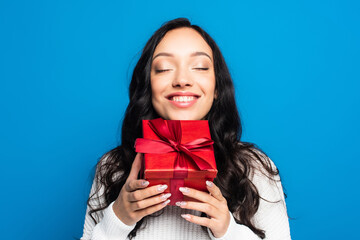 Fototapeta premium happy brunette woman with closed eyes holding christmas present isolated on blue