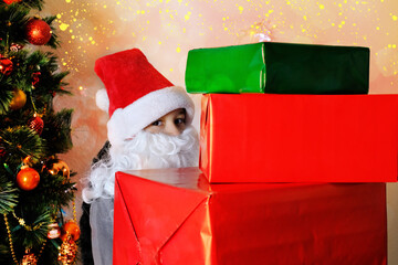 little kid, santa claus helper, an elf with a white beard holds gifts near the christmas tree, the...
