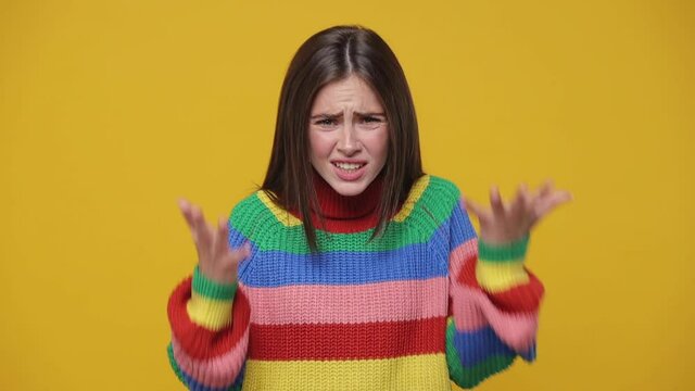 Shocked perplexed angry irritated young woman 20s years old in colorful rainbow sweater posing isolated on yellow background studio. People lifestyle concept. Looking camera spreading hands swearing