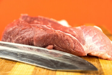 Close-up on a cutting board of a large worker's knife and a piece of raw fresh meat on a bright orange background. Selective focus, shallow depth of field