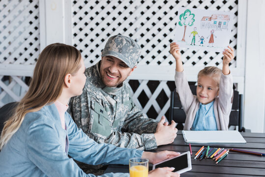 Smiling man in military uniform looking at woman with digital tablet, while sitting with daughter showing picture at home