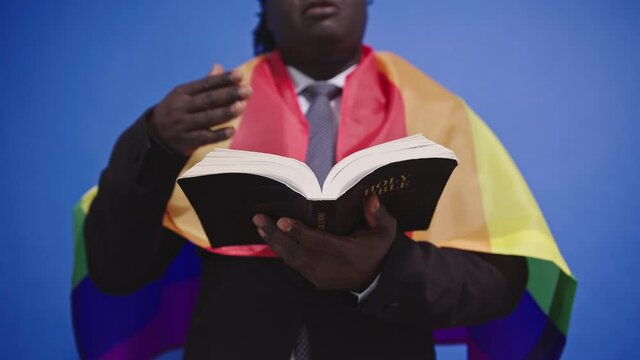 Black Man Caped With a Rainbow LGBT Flag Reading Holy Bible, Close Up. Religious African Priest Supporting Gay Community, Slow Motion